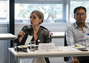 María Eugenia Lago, directora general del IDECBA, hablando en un micrófono durante su participación en el Programa Ejecutivo de Alto Nivel del PNUD en Lima, Perú. Se la ve sentada en una mesa blanca, con un cartel identificatorio con su nombre y cargo. A su lado derecho, se observa a otro participante del evento.