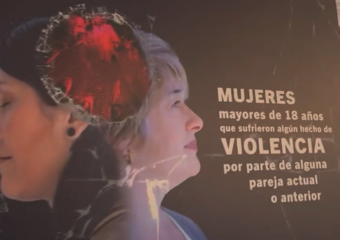 Primer plano de un cartel de la muestra fotográfica 'Un fondo negro', exhibido en la Facultad de Ciencias Económicas de la UBA. El cartel muestra el perfil de dos mujeres y un texto que dice 'MUJERES mayores de 18 años que sufrieron algún hecho de VIOLENCIA por parte de alguna pareja actual o anterior', destacando la frase 'VIOLENCIA' en mayúsculas y negrita, con un efecto de vidrio roto sobre la cabeza de una de las mujeres, simbolizando el impacto de la violencia de género.