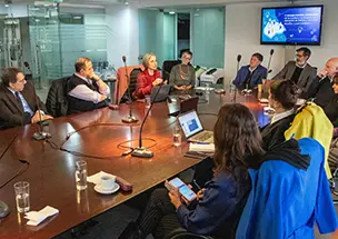 Mesa de trabajo en el Consejo Económico y Social de la Ciudad de Buenos Aires con participantes debatiendo sobre el envejecimiento poblacional. En la imagen se observa a varias personas sentadas alrededor de una mesa de reuniones de madera, con María Eugenia Lago, directora general del IDECBA, participando activamente. Al fondo, una pantalla muestra gráficos relacionados con el tema