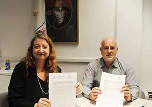 El director ejecutivo del IDECBA, José María Donati, y la defensora del pueblo de la Ciudad de Buenos Aires, María Rosa Muiños, posando con los documentos del convenio de cooperación firmados. Ambos están de pie, sonriendo, con un retrato y una bandera institucional de fondo